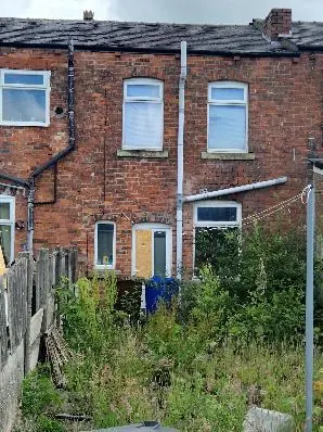Rear exterior of a brick terraced property with an overgrown garden and boarded window
